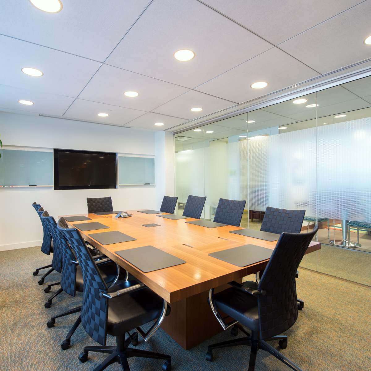 A modern conference room features a large wooden table surrounded by black chairs, with a glass wall and a wall-mounted screen.