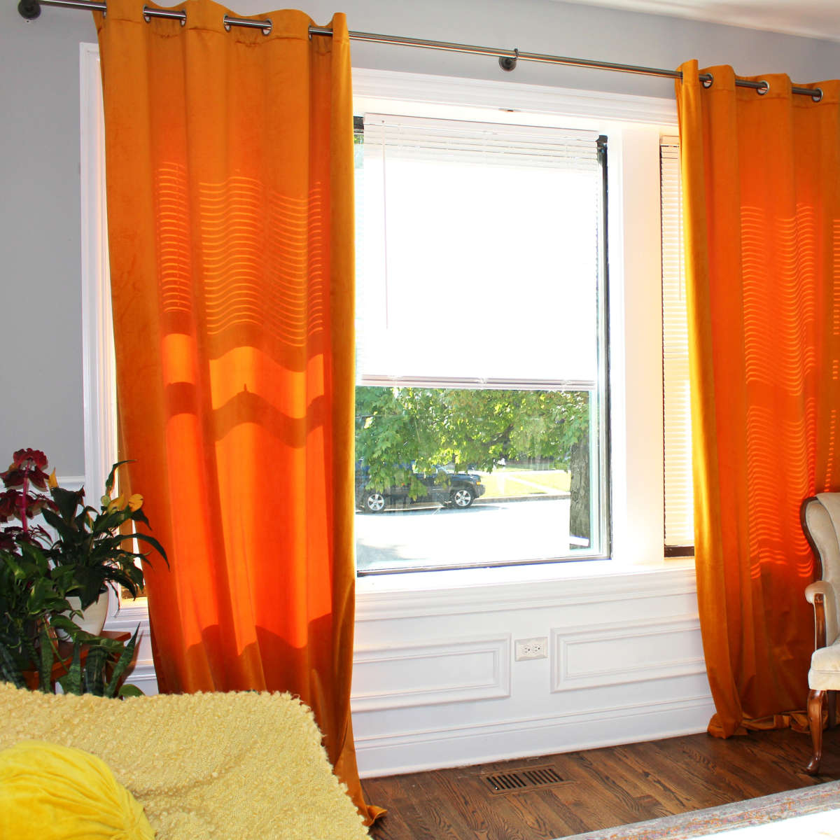 A cozy living room with orange curtains framing a window, a floral painting on the wall, a small armchair, and plants nearby.