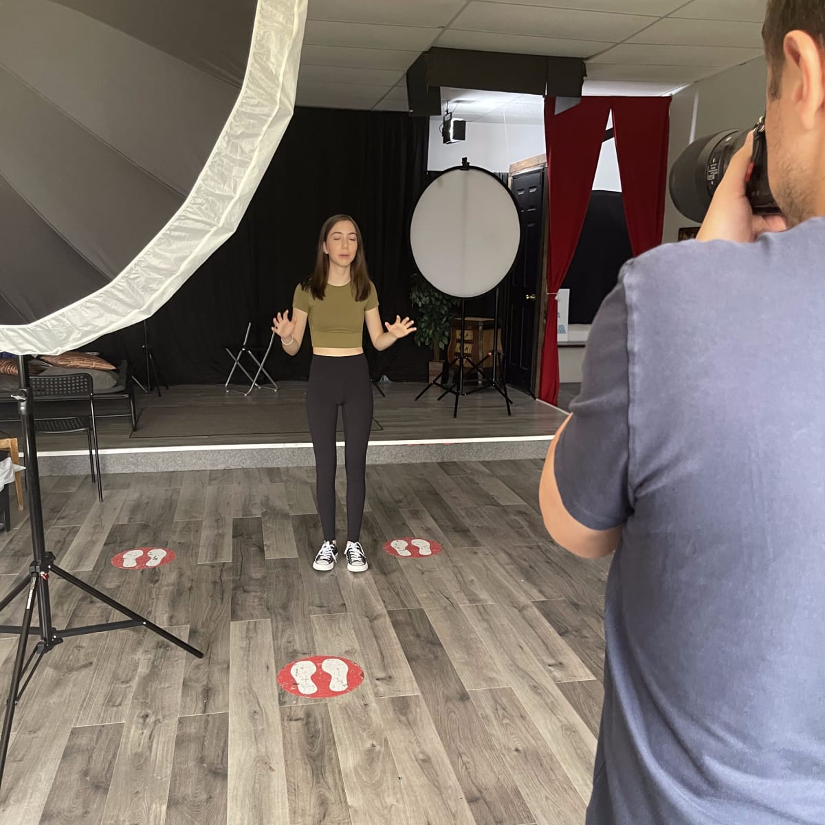 A young woman stands in a photography studio, while a person behind a camera prepares to take her picture.