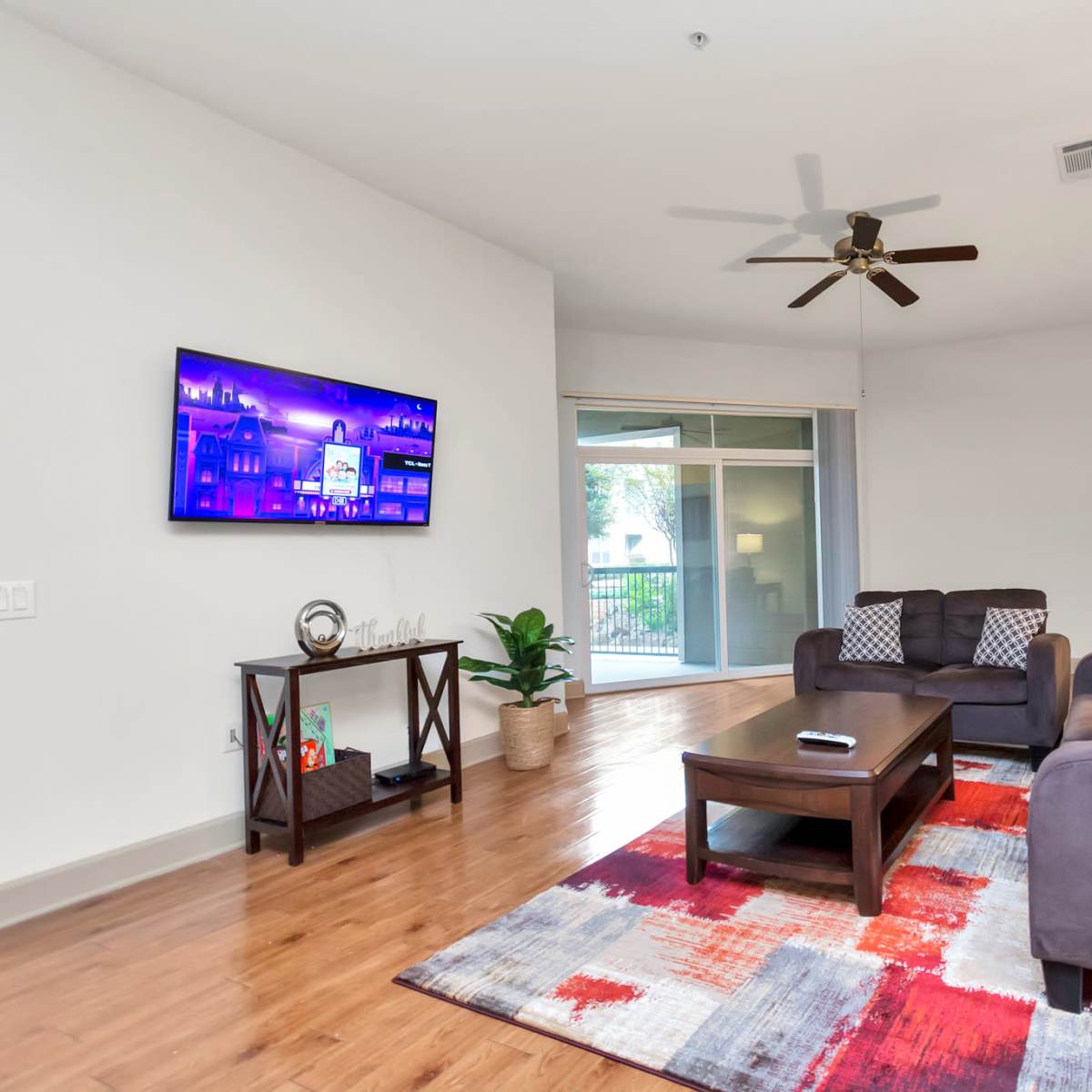 The living room features a dark gray sofa and an armchair arranged around a coffee table, with a large television mounted on the wall and a sliding glass door leading to a balcony.