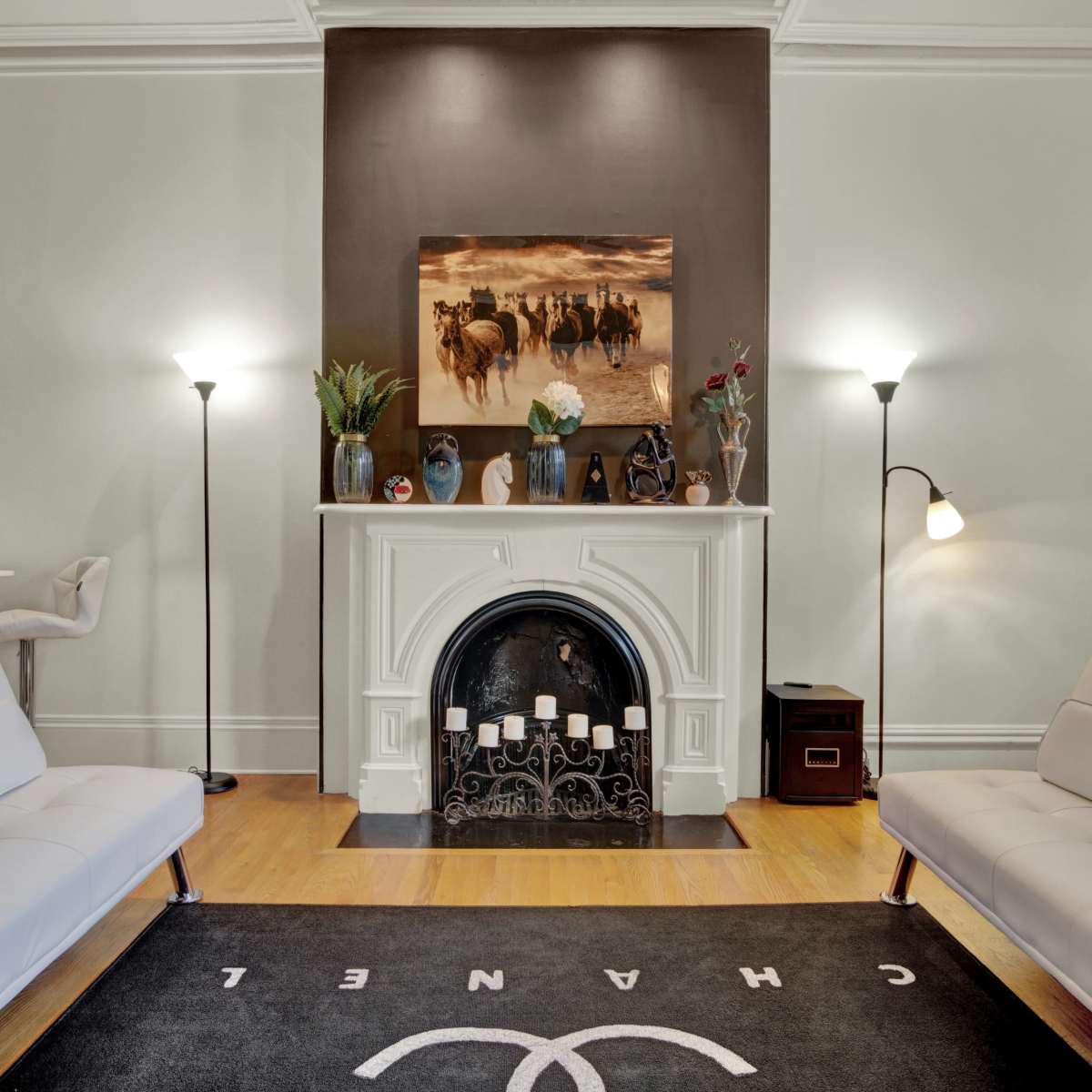 A living room featuring a fireplace with a decorative painting above it, flanked by two lamps, a carpet with a logo, and two white sofas facing each other.