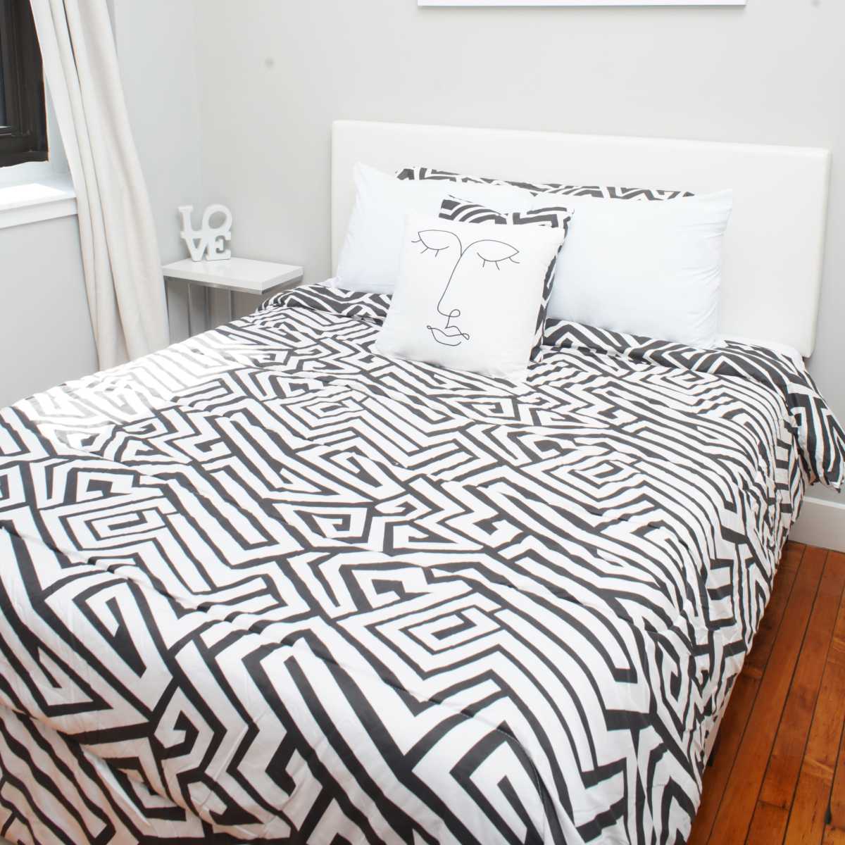 A neatly made bed with a black and white patterned duvet and a decorative pillow featuring a minimalistic face design is positioned against a light-colored wall, beneath a framed sign that reads "NAMASTAY in bed."