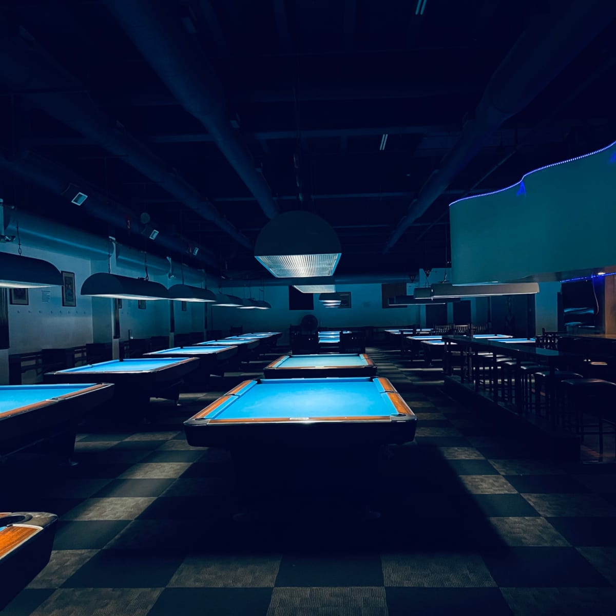 An empty pool hall features several tables, each illuminated by overhead lights, under dim conditions.