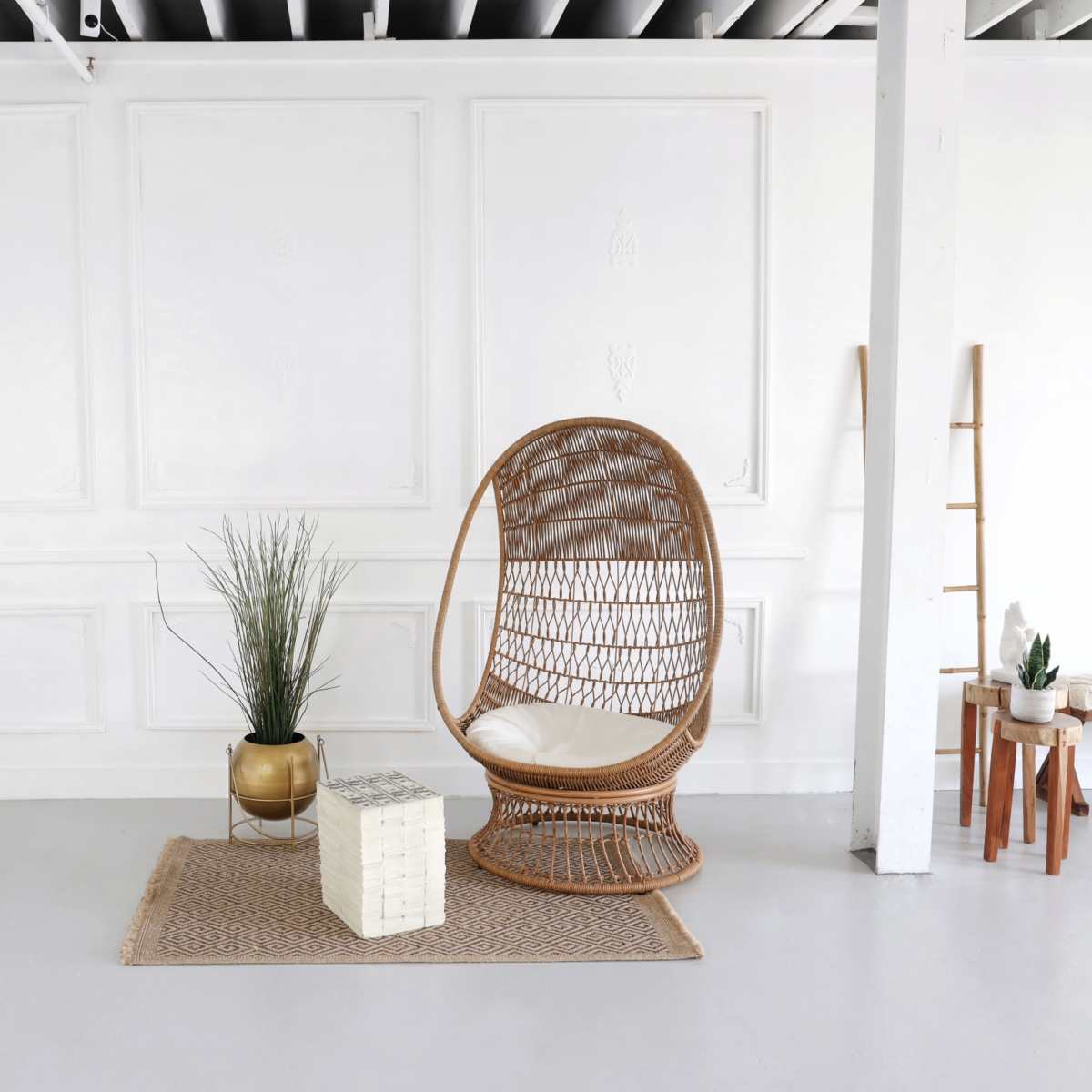 A minimalistic interior space featuring a woven rattan chair, a round gold planter with greenery, a decorative woven rug, a tall mirror, and a small side table.