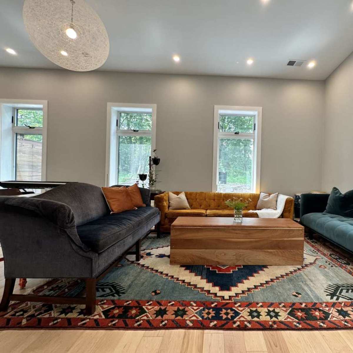 A well-furnished living room features a mix of seating options, including a gray sofa, a yellow tufted couch, and a blue-green sectional, arranged around a wooden coffee table on a patterned area rug.