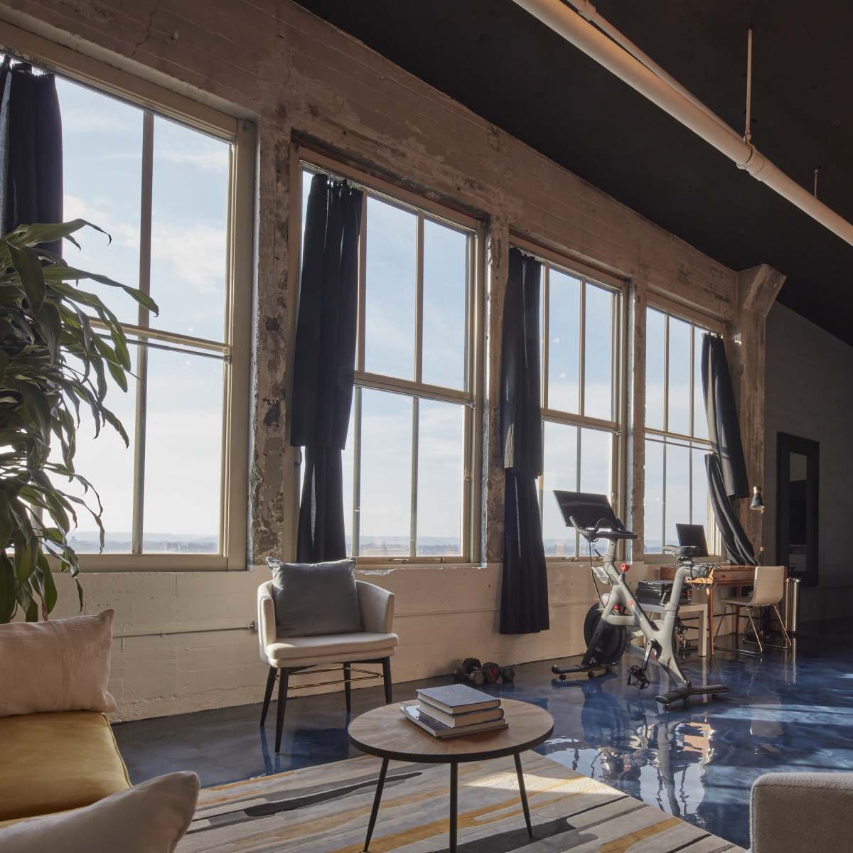 An industrial-style living space with large windows, a workout bike, a kitchen area, and modern furniture.