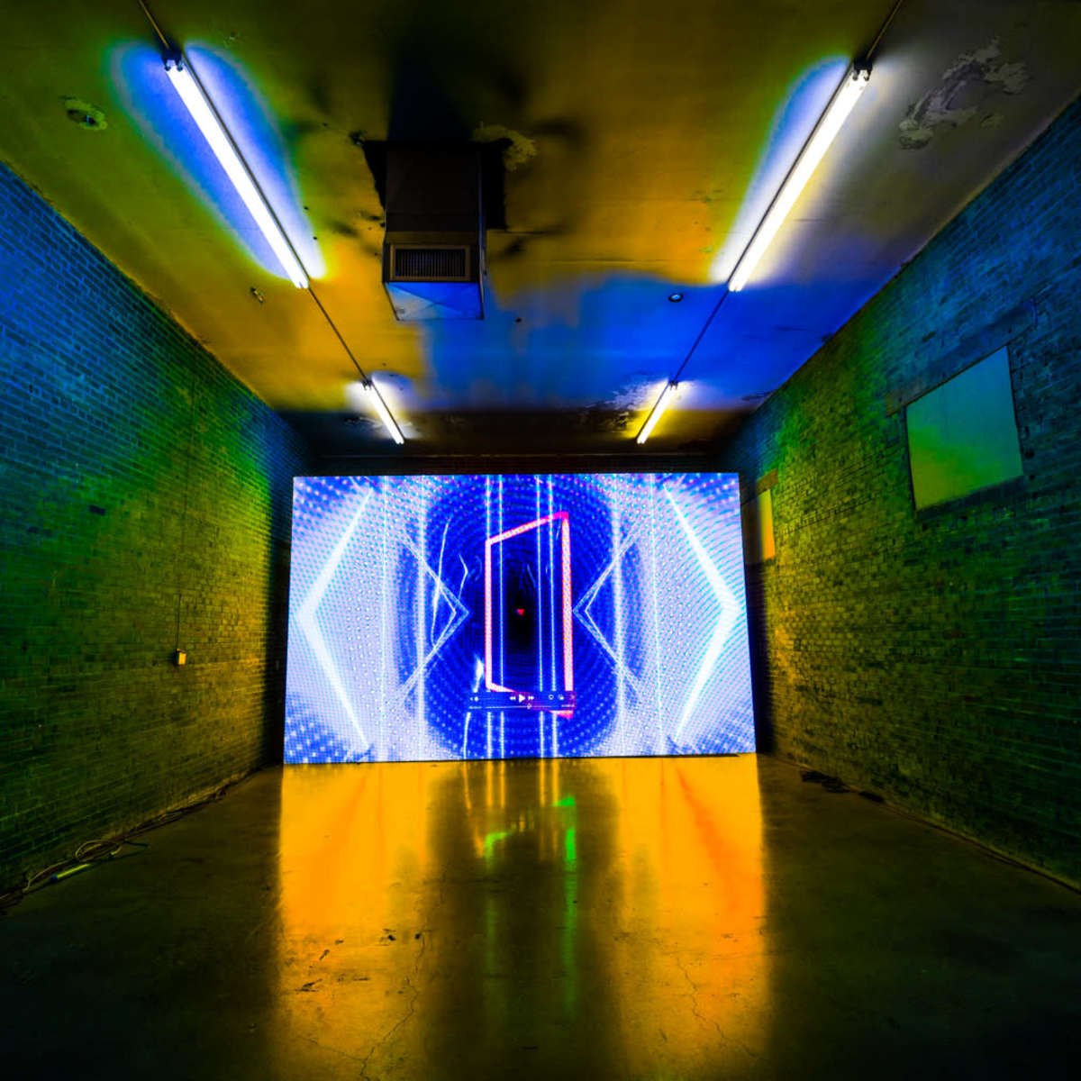 A dimly lit room features a large, brightly illuminated screen displaying dynamic visual patterns on the far wall.