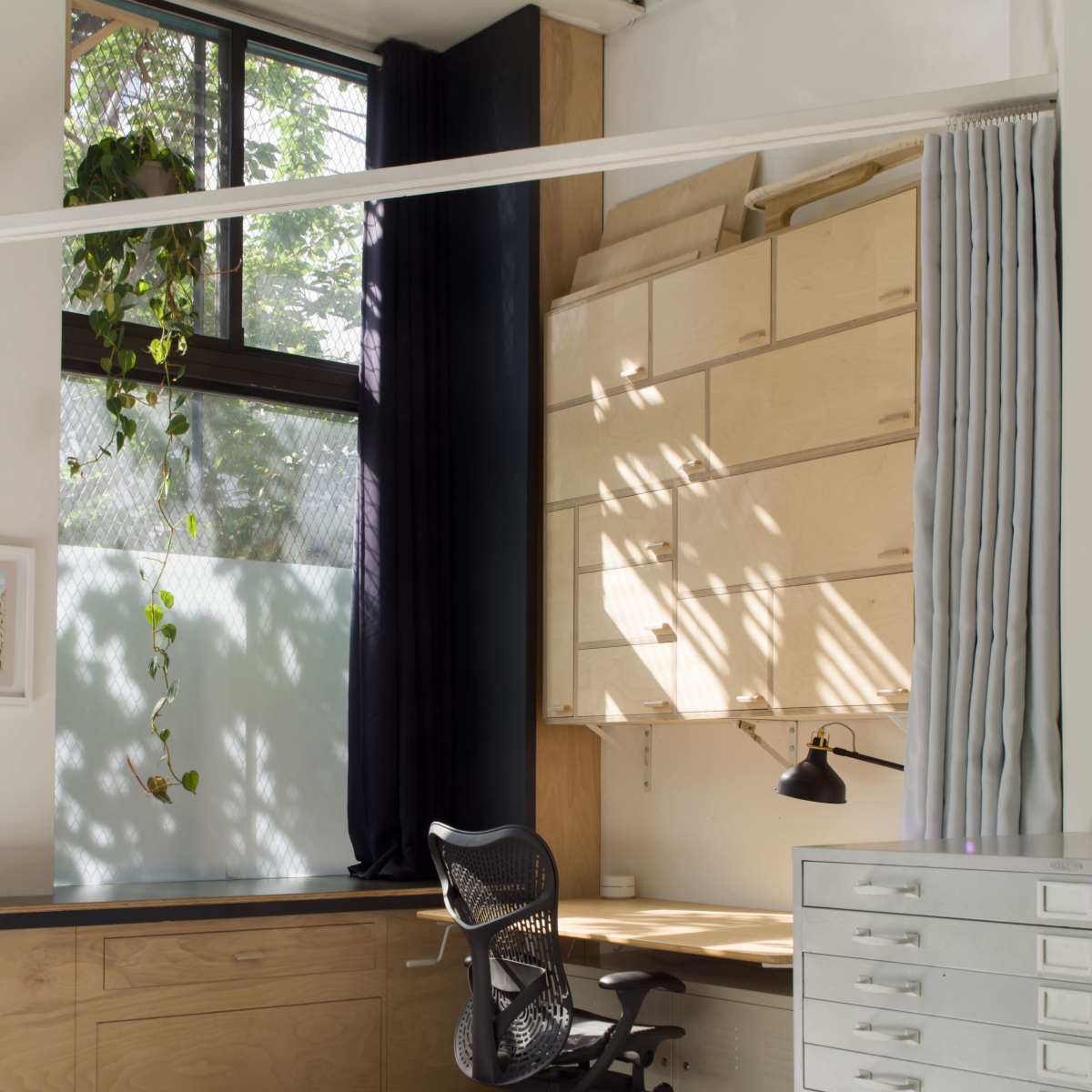 A minimalist home office with a wooden desk and chair, wall-mounted storage, and natural light filtering through a large window.