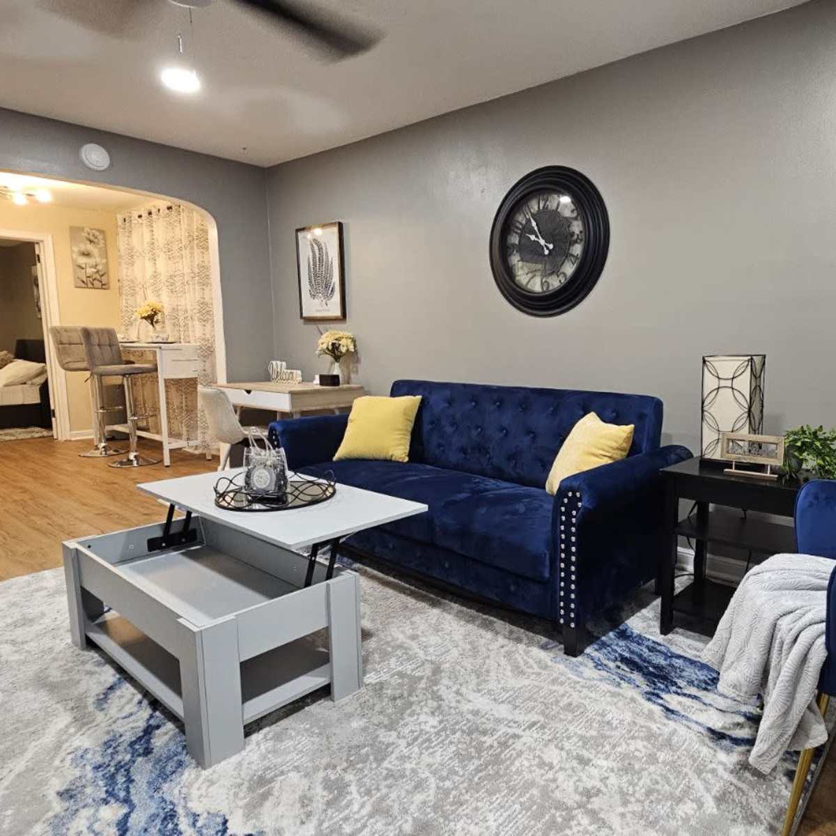 A modern living room featuring a blue velvet sofa, a light gray coffee table, a decorative plant, and a prominent wall clock.