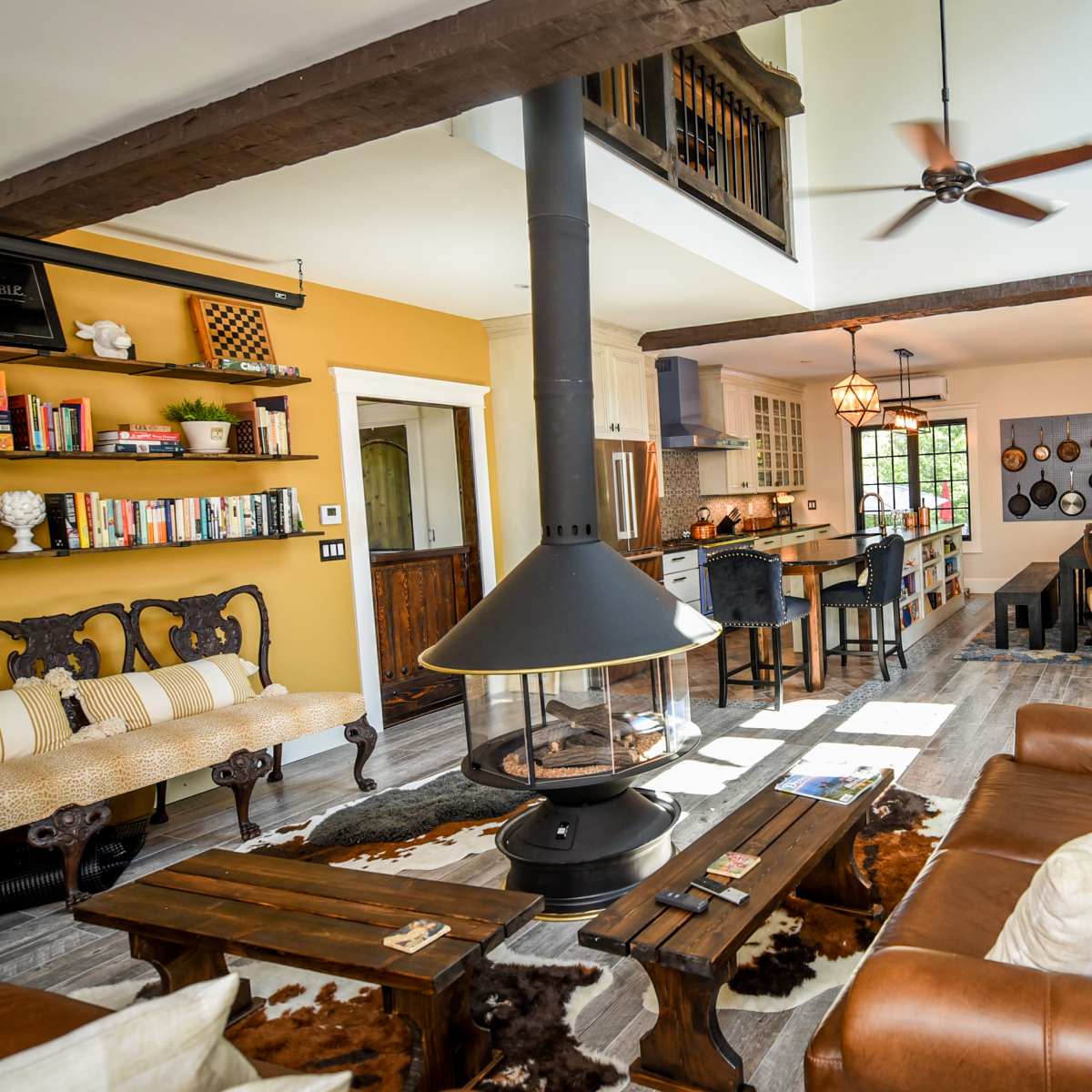 A living area features a brown leather sofa, a central fireplace, and an open layout connecting a dining space and kitchen with wooden beams and warm-colored walls.