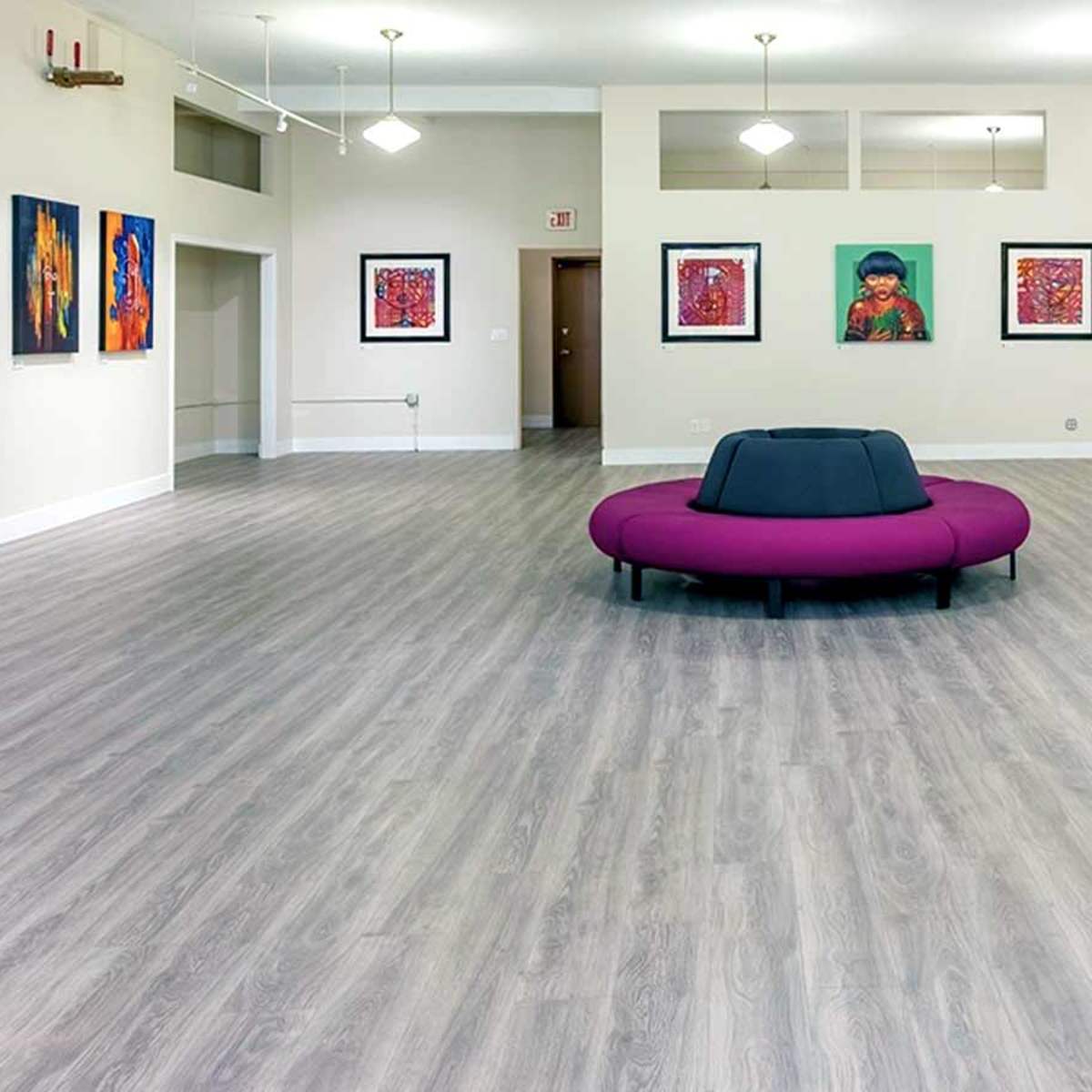A brightly lit gallery space with a large purple seating area in the center and colorful artworks displayed on the walls.