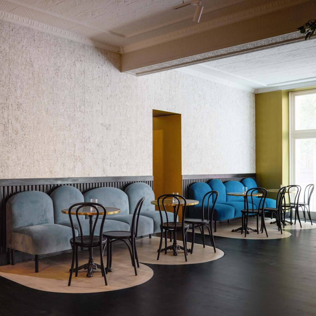 A café interior with two rows of seating and tables, featuring black chairs and plush blue and gray couches against textured walls.