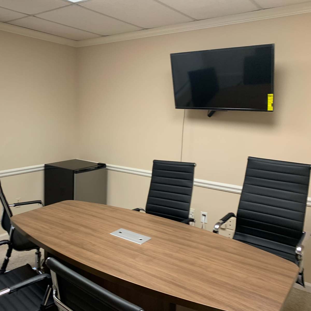 A conference room features a large wooden table, six black office chairs, a flat-screen TV mounted on the wall, and a small refrigerator in the corner.