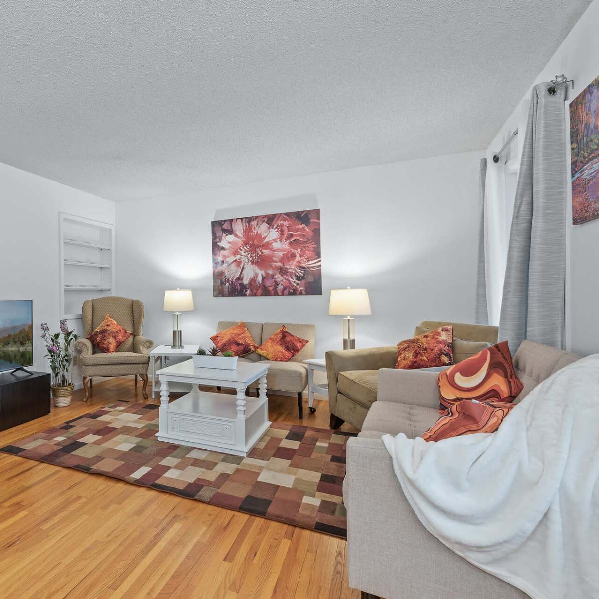 The living room features a beige sofa, two armchairs, a coffee table, and a television placed on a cabinet, all arranged around a colorful area rug.