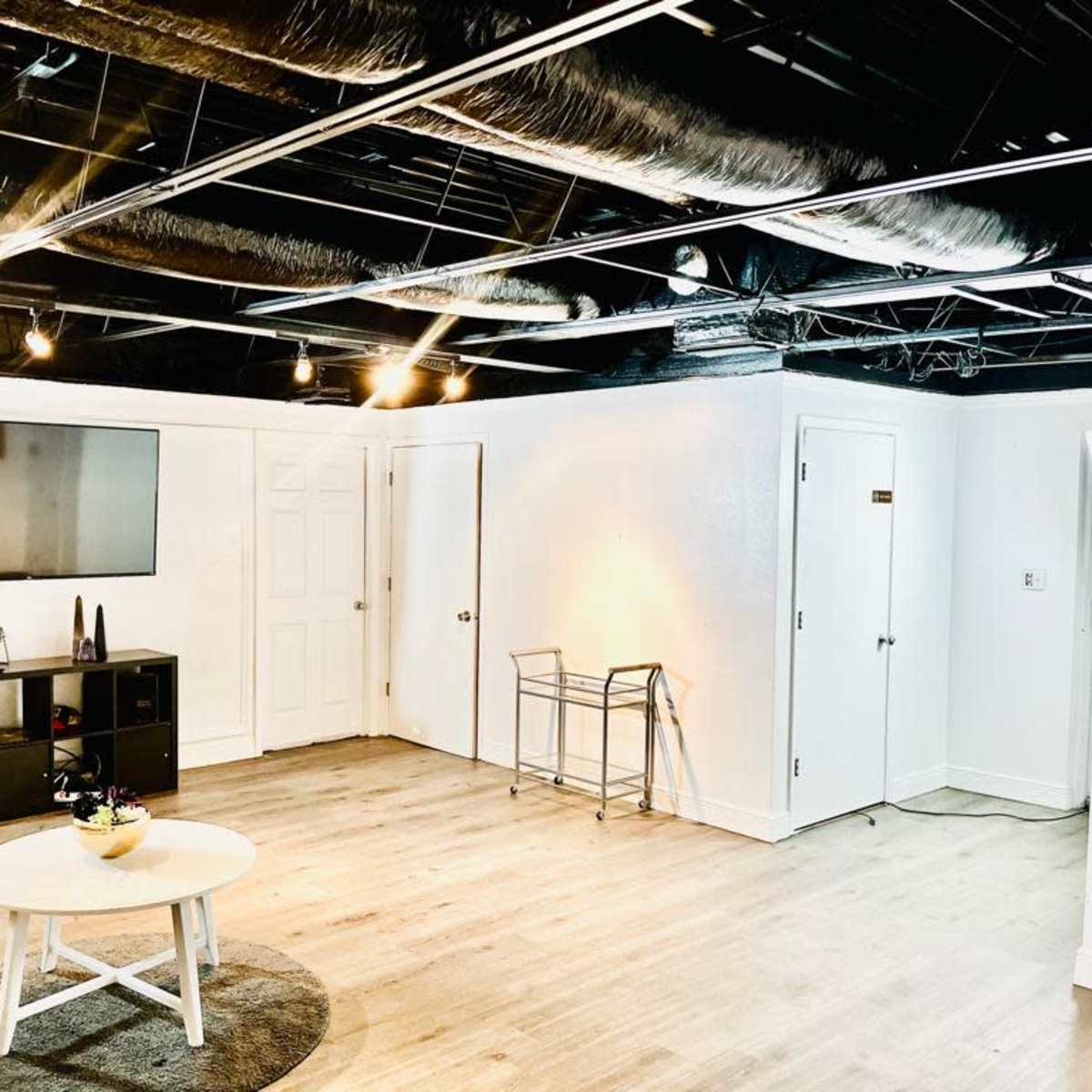 A spacious, well-lit room with a sofa, a coffee table, a flat-screen TV mounted on the wall, and several doors leading to other areas.
