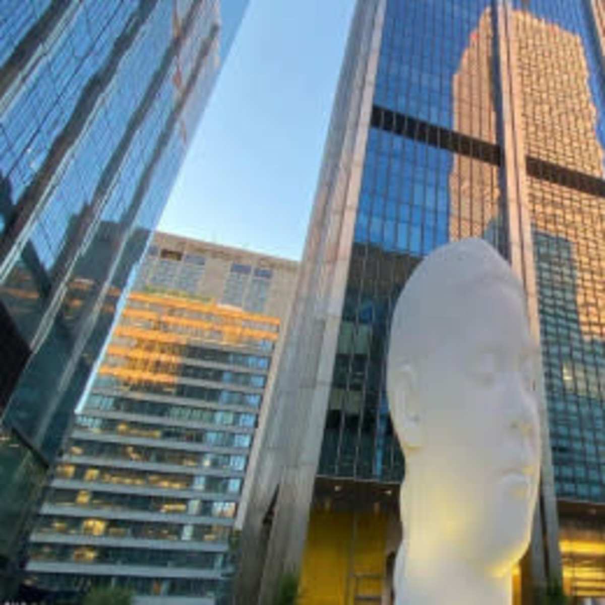 A large white sculpture of a head stands in front of reflective glass skyscrapers.