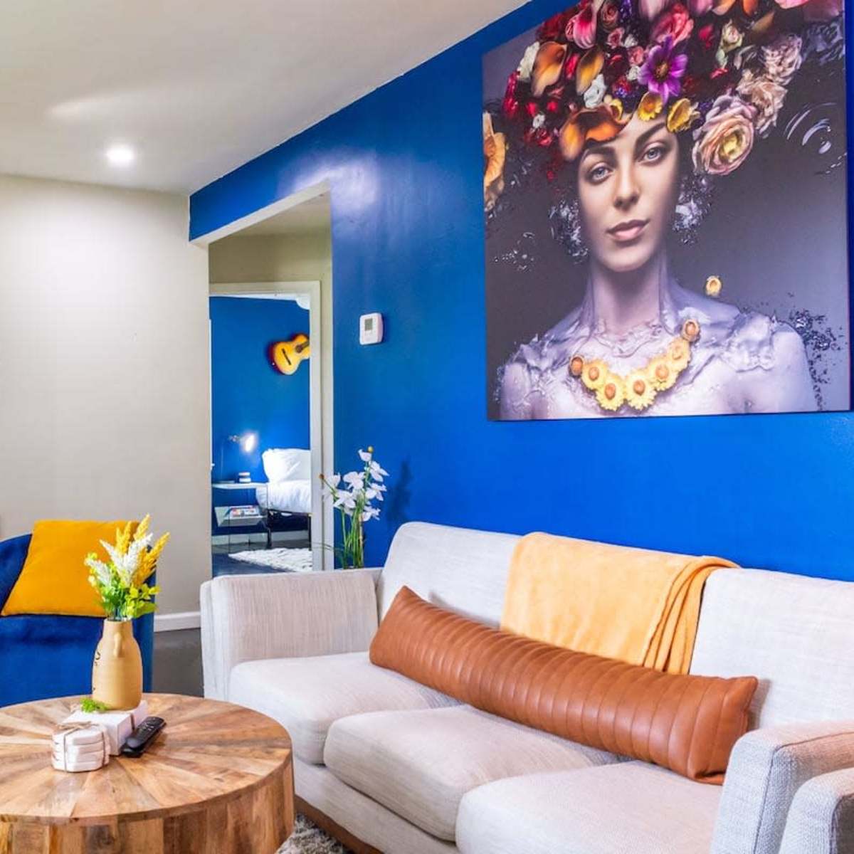 A brightly colored living room features a circular wooden coffee table, a gray sofa with an orange throw, and blue chairs with yellow pillows, alongside a large portrait on the wall.