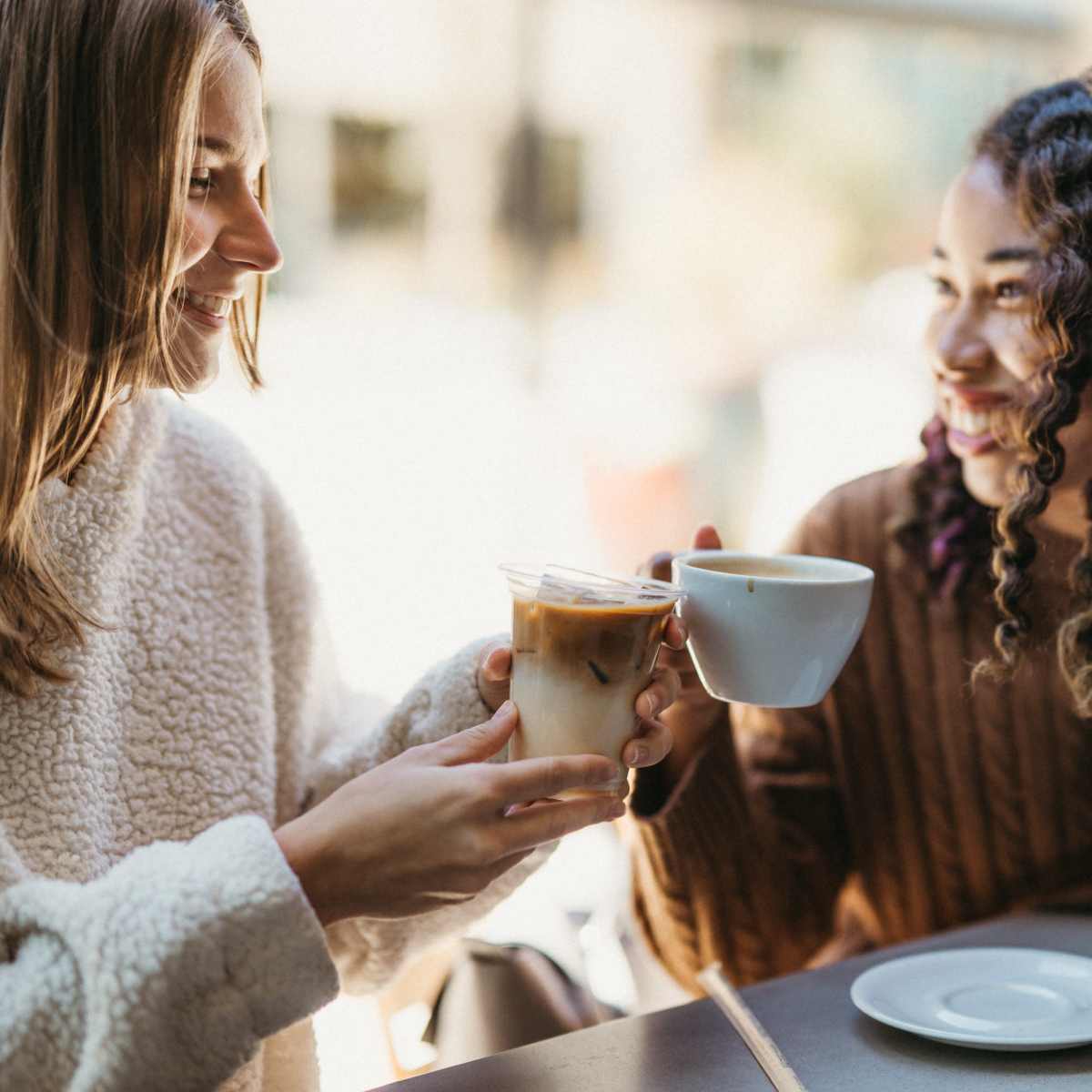 Two women sit at a table and toast their drinks in a bright cafe.