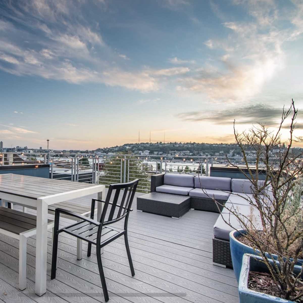 A rooftop terrace features a white dining table and black chairs, overlooking a city skyline and dotted with potted plants.