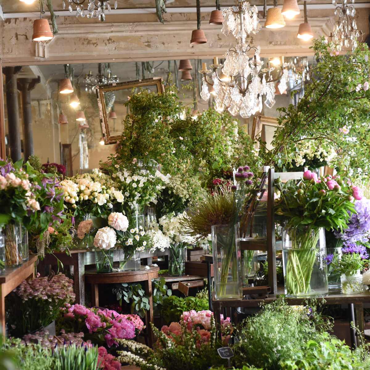 A floral shop filled with a variety of colorful flowers arranged in glass vases on wooden tables, under a chandelier and surrounded by mirrors.