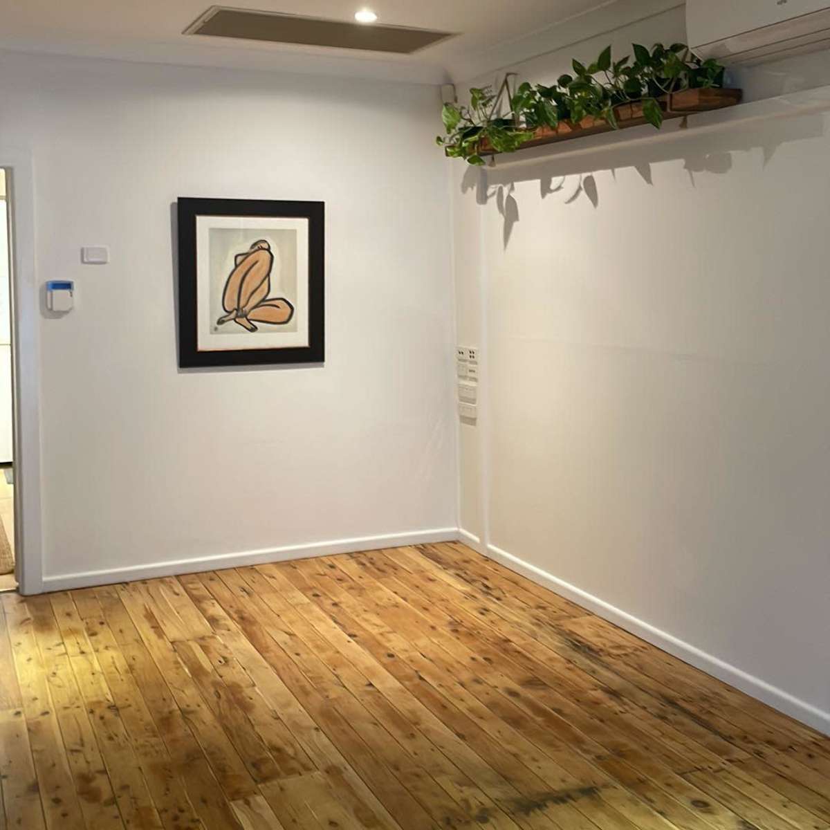 A minimalistic room with wooden flooring, a framed artwork on the wall, and a shelf with plants above it.