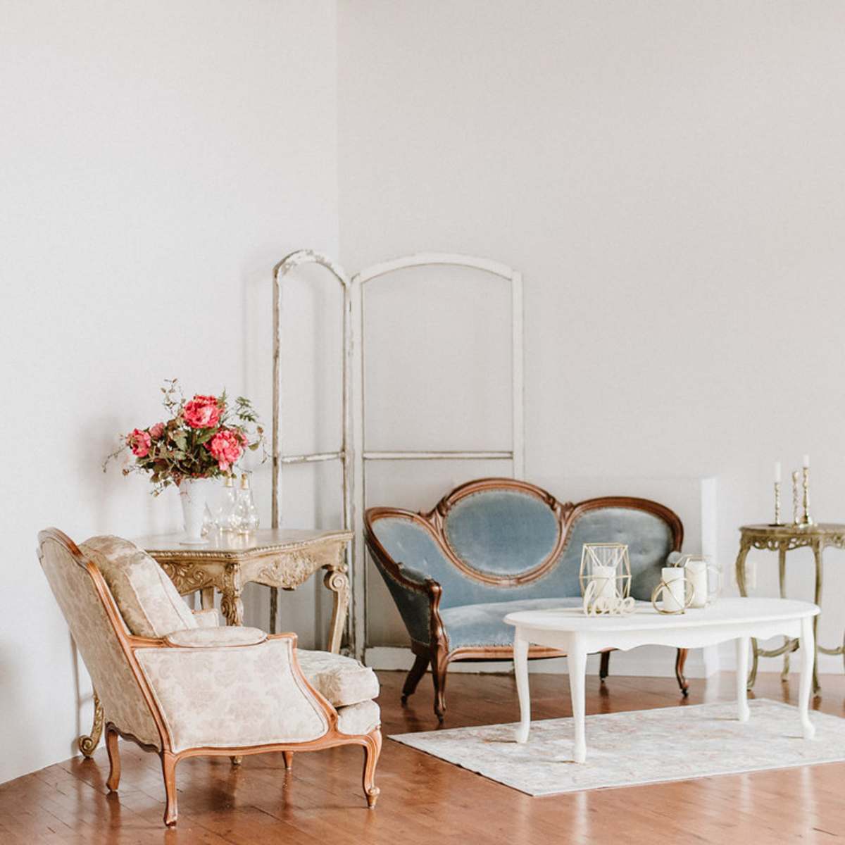 A softly lit room featuring vintage furniture, including a floral sofa, an upholstered chair, and a white coffee table, arranged on a wooden floor.