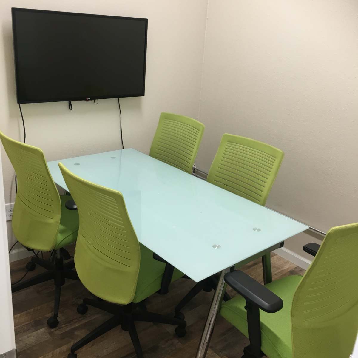 A small conference room features a glass table surrounded by five green office chairs and a wall-mounted television.