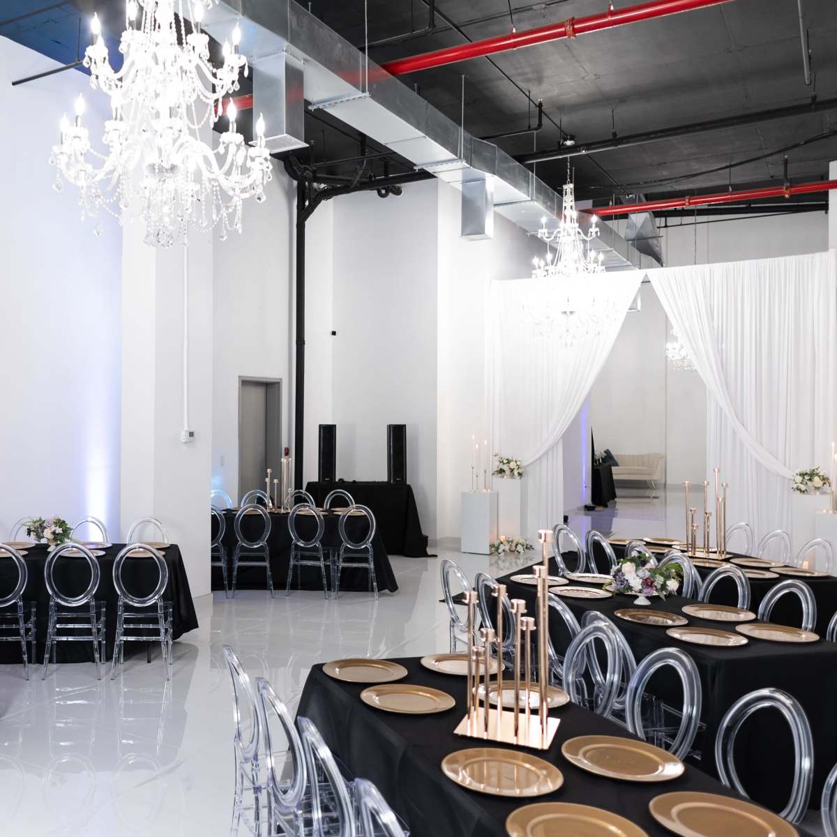 A spacious event hall features black tables set with gold chargers and illuminated by chandeliers.