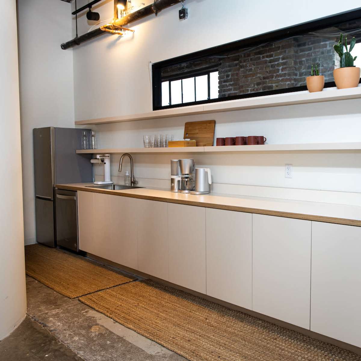 A minimalist kitchen with a long white countertop, shelves displaying dishes and a plant, and a stainless steel refrigerator.