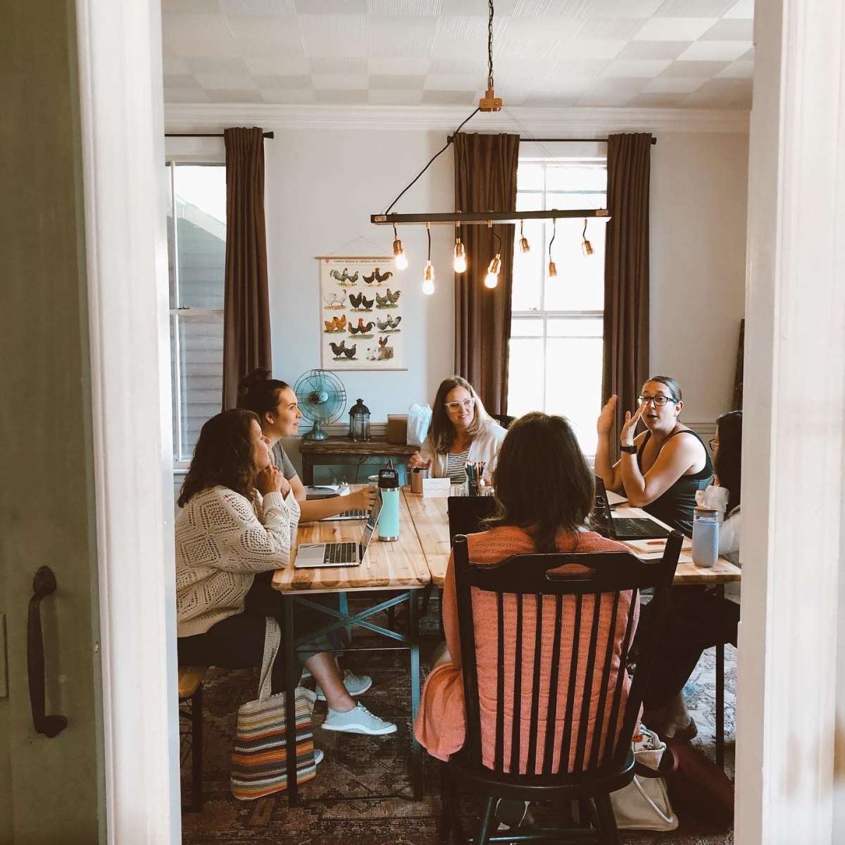A group of six people sits around a wooden table in a well-lit room, engaged in a discussion with laptops and water bottles in front of them.