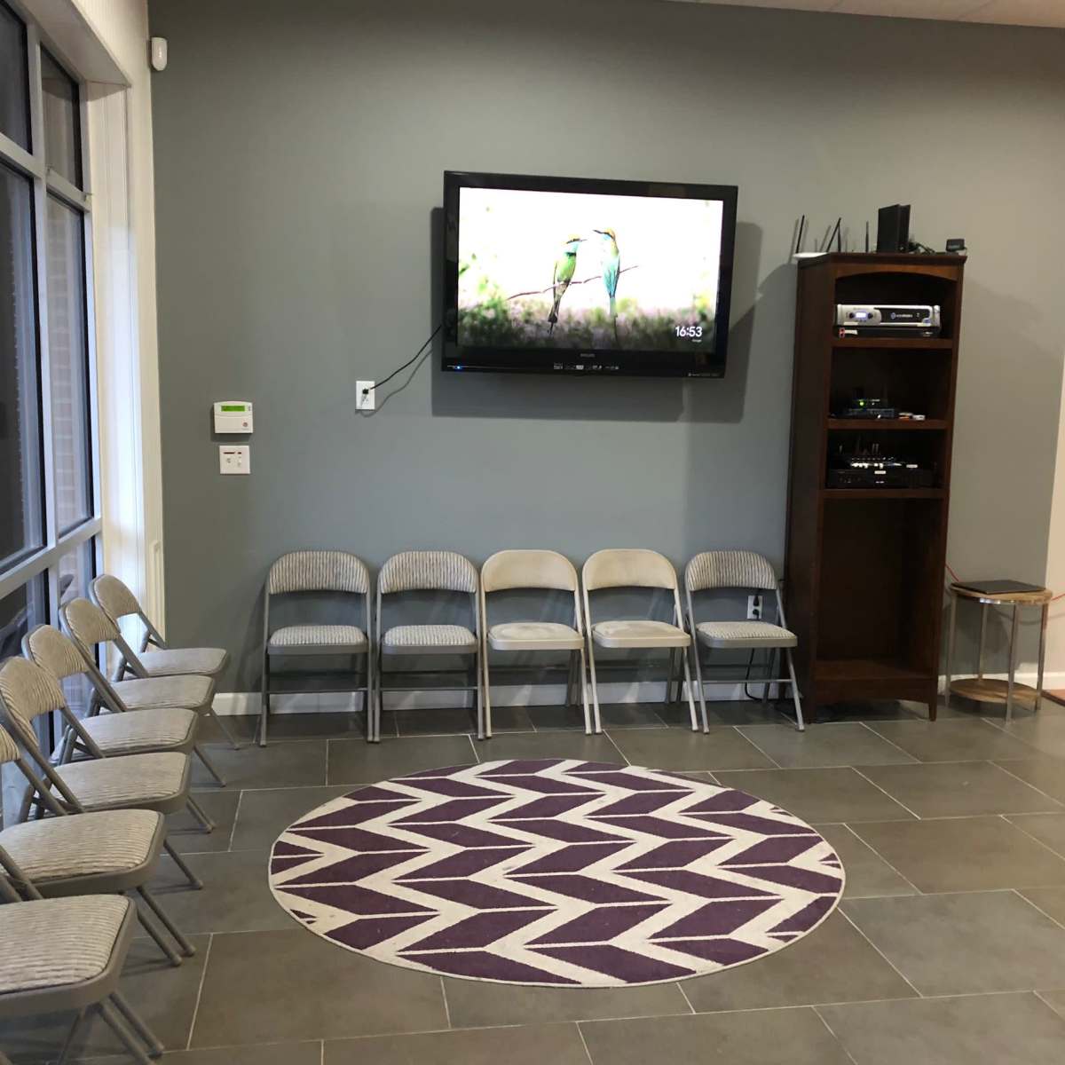 A waiting area features six metal folding chairs arranged in a semi-circle around a purple and white patterned rug, with a wall-mounted TV and a wooden shelving unit in the background.