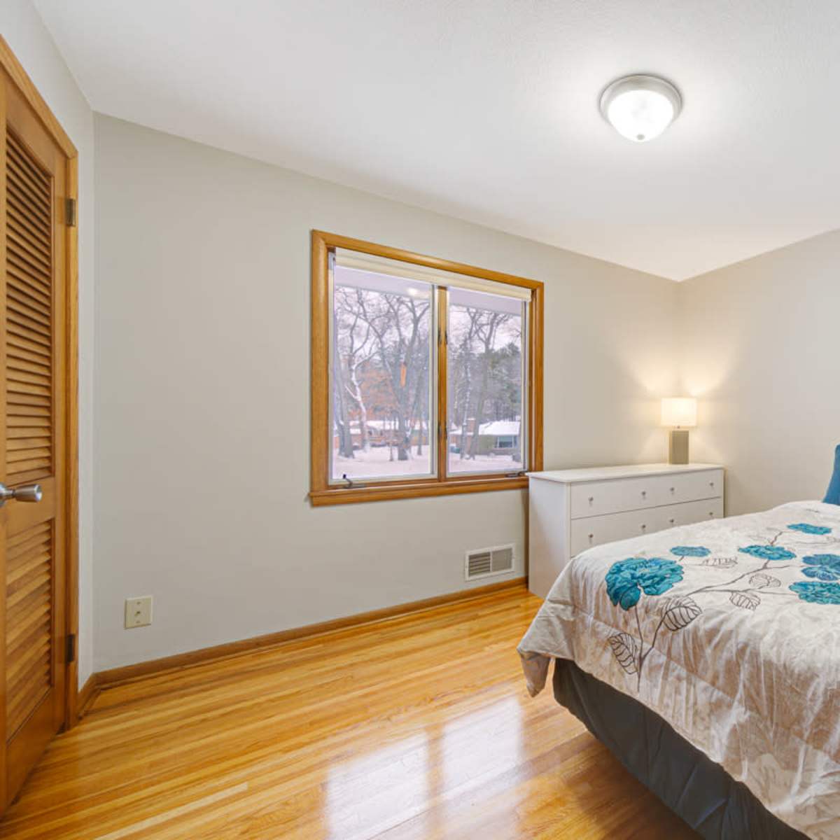 A well-lit bedroom features a bed with a floral-patterned comforter, a dresser, and a window overlooking a snowy landscape.