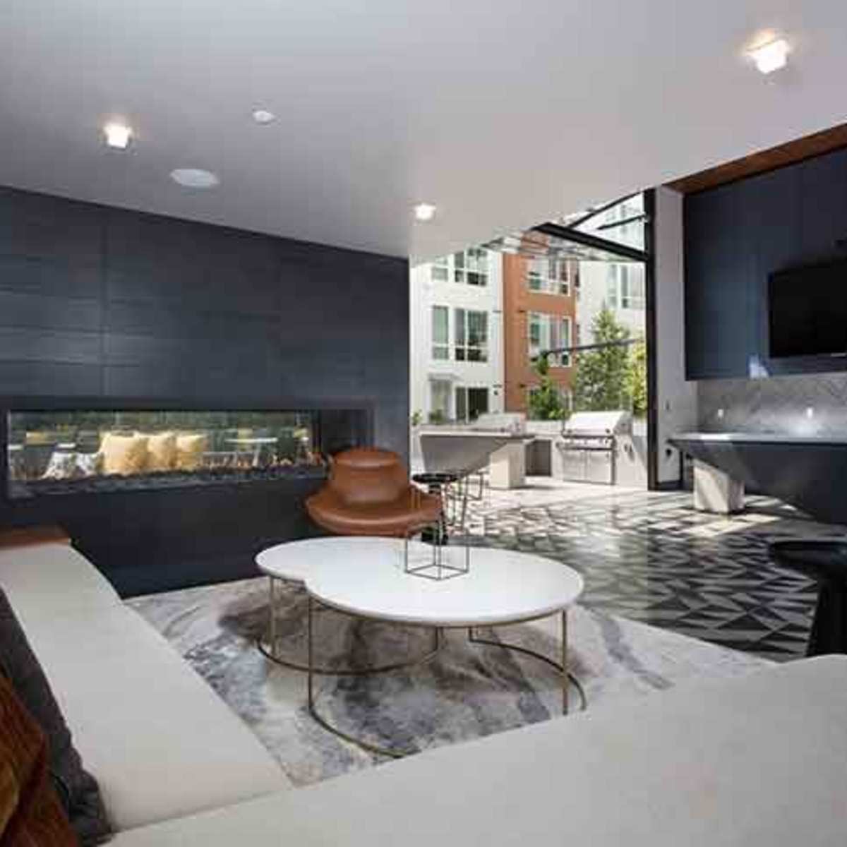 A modern living area featuring a sectional sofa, a coffee table on a patterned rug, a wall-mounted television, and large windows overlooking an outdoor space.