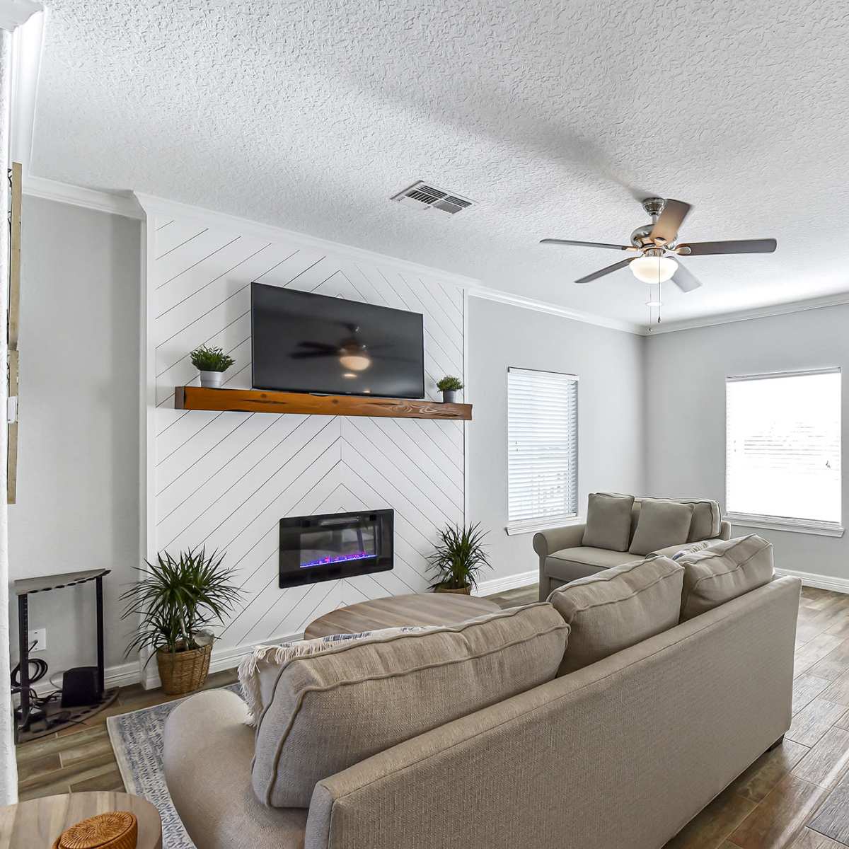 A modern living room with a light-colored sofa, a wall-mounted TV above a fireplace, and large windows letting in natural light.