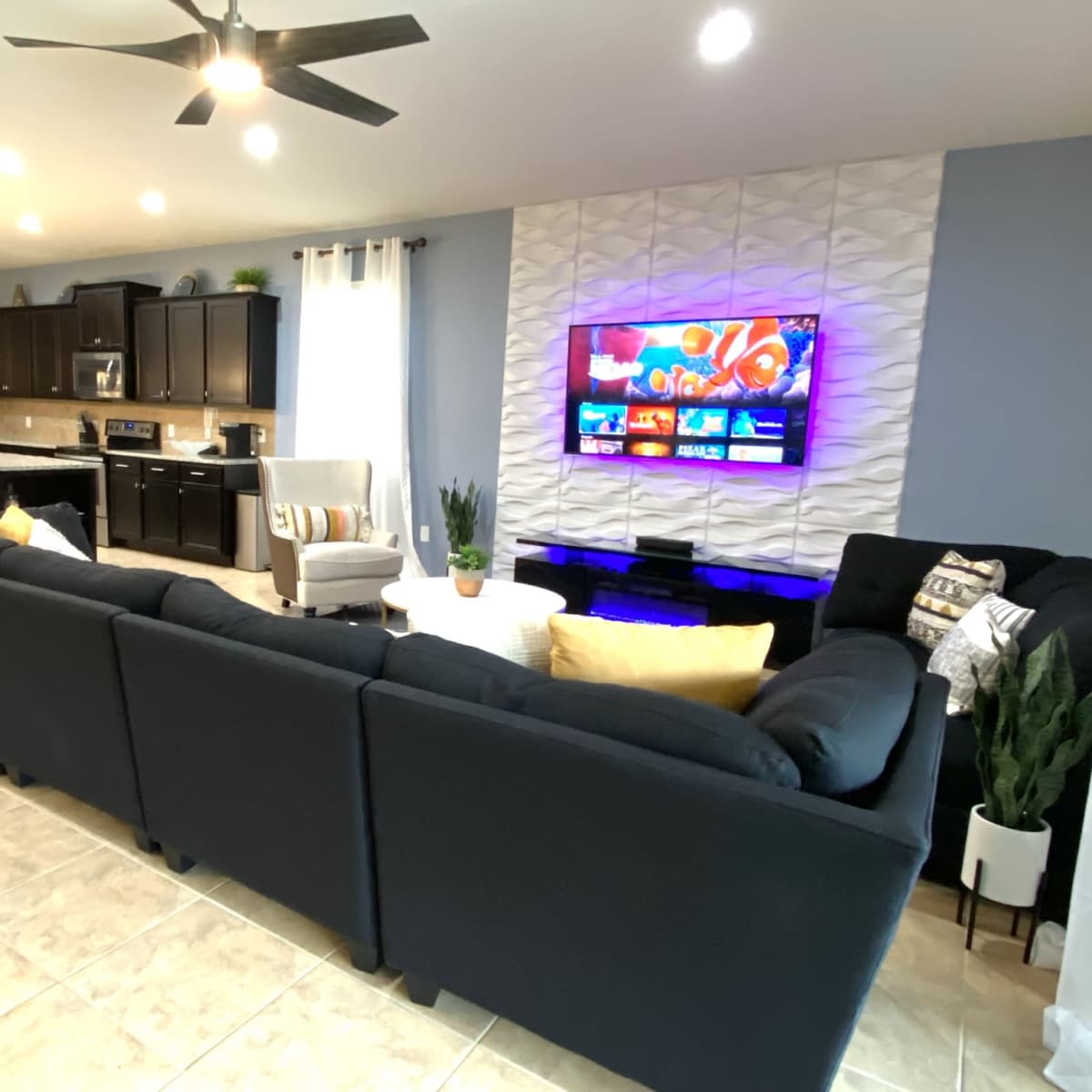 A modern living room with a sectional sofa facing a wall-mounted television, adjacent to a kitchen with dark cabinetry and bar seating.