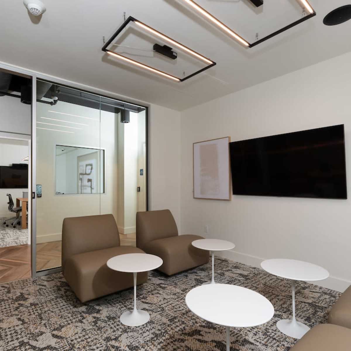 A modern meeting room with two brown chairs, white coffee tables, and a large flat-screen TV mounted on the wall.
