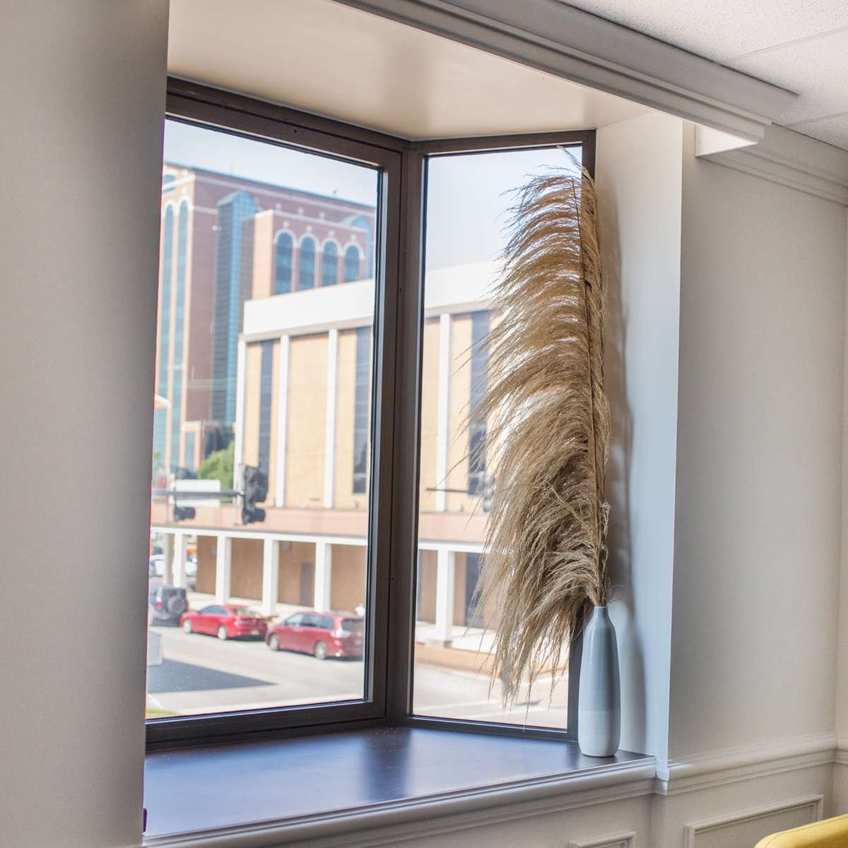 A bright corner window features a tall, dried grass arrangement in a vase, with a view of a city street and buildings outside.