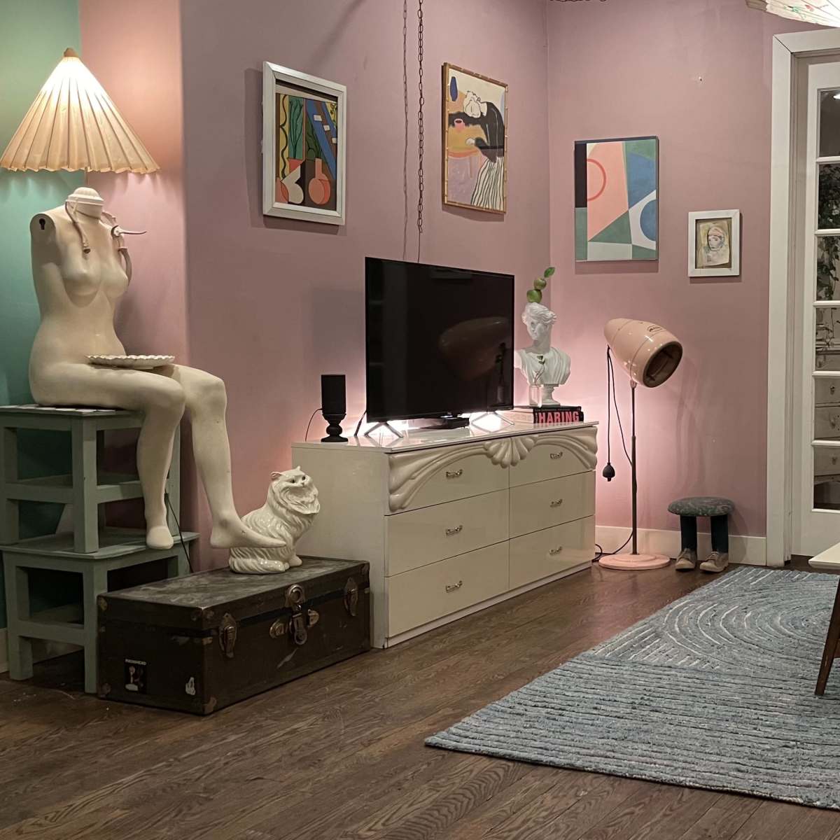 A cozy living room with pastel pink walls, featuring a television, a mannequin, various artwork, and a mix of furniture including a green couch, coffee table, and a standing lamp.