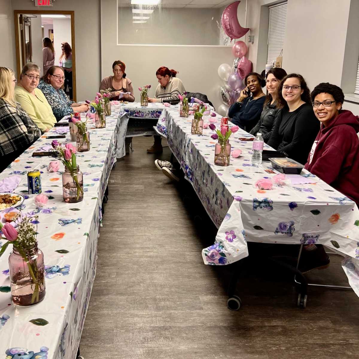 A group of people is seated at a long, decorated table with floral tablecloths, enjoying a gathering in a bright, indoor setting.