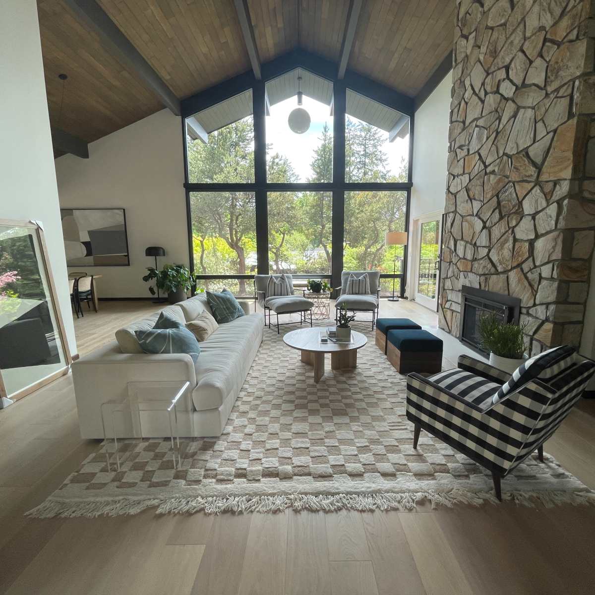 The scene features a spacious living room with a large stone fireplace, a mix of modern and patterned furniture, and large windows overlooking a garden.