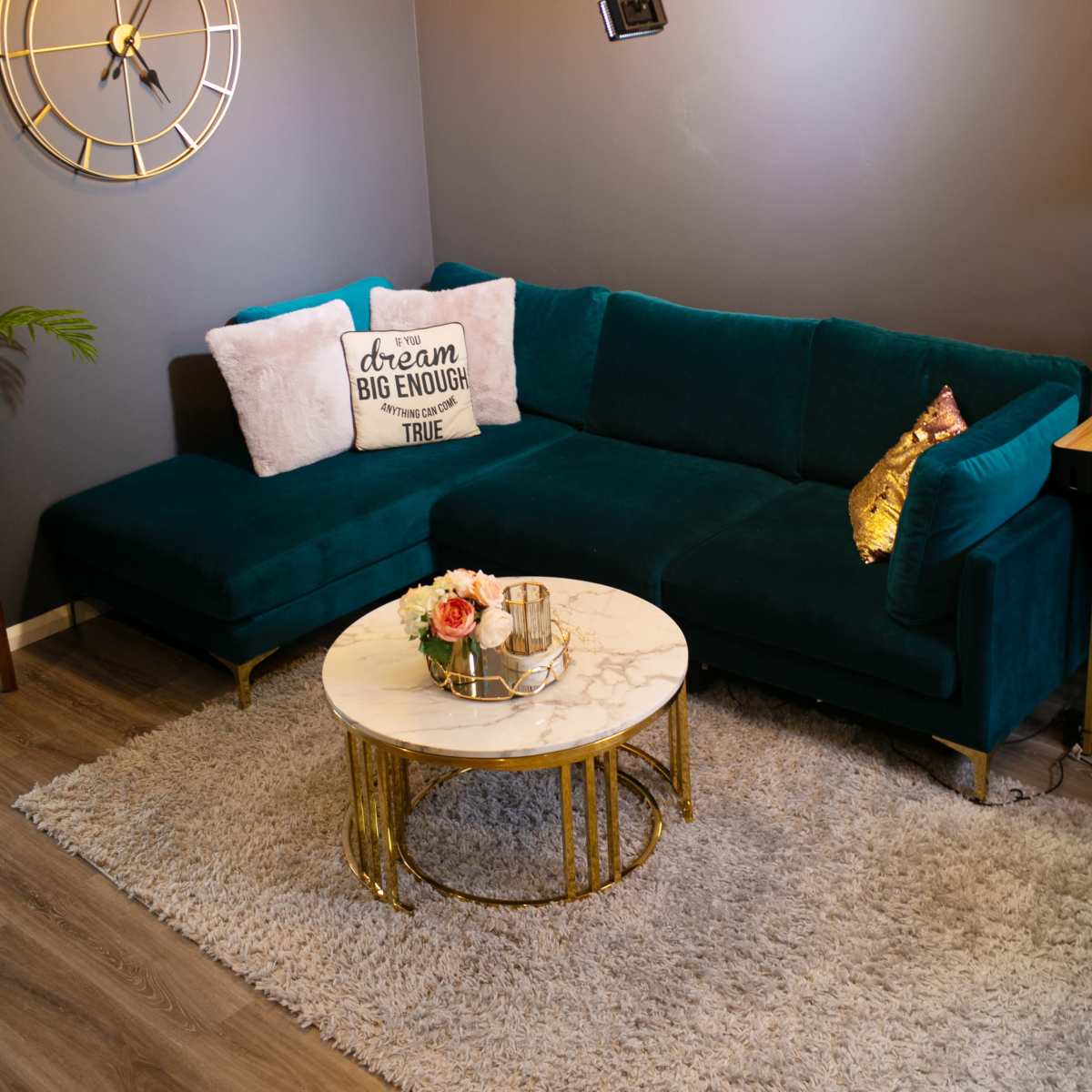 A modern living room setup with a teal sectional sofa, a round marble coffee table, and decorative pillows, illuminated by pendant lights and a wall-mounted spotlight.