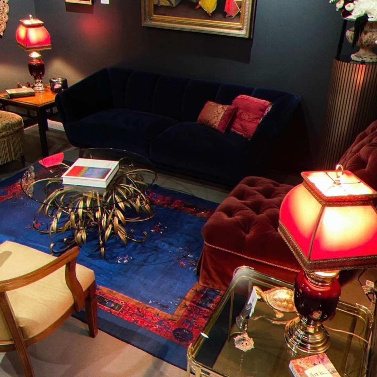 A stylish sitting area featuring a dark blue velvet couch, a red tufted chair, a decorative rug, and various decorative items, including lamps and artwork on the walls.