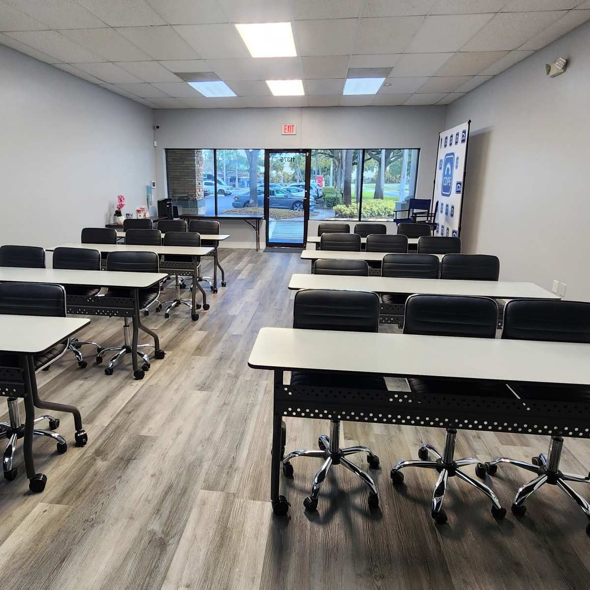 A brightly lit classroom with rows of steel-frame tables and black rolling chairs facing the entrance.