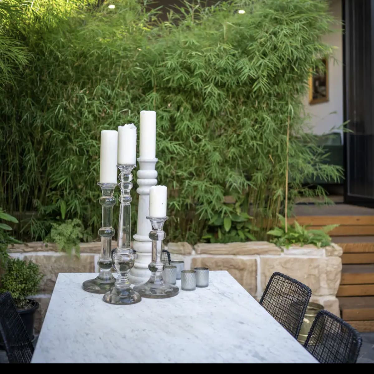 A marble table with candle holders is situated in a garden surrounded by bamboo, leading up to a doorway with wooden steps.