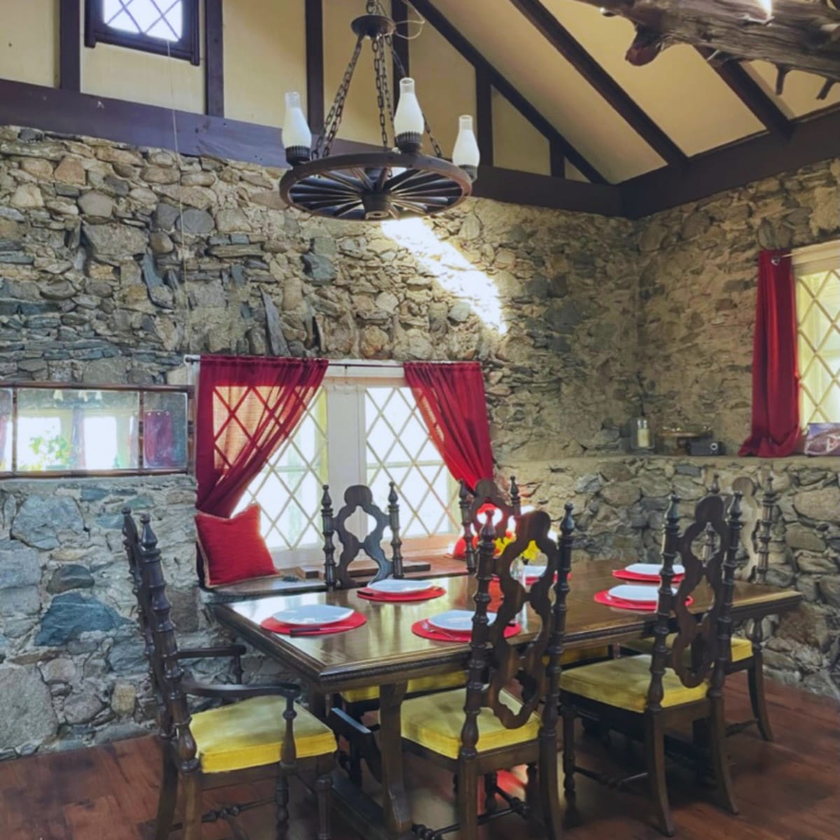 A rustic dining area features a wooden table surrounded by high-backed chairs, set against a stone wall with red curtains framing the windows.