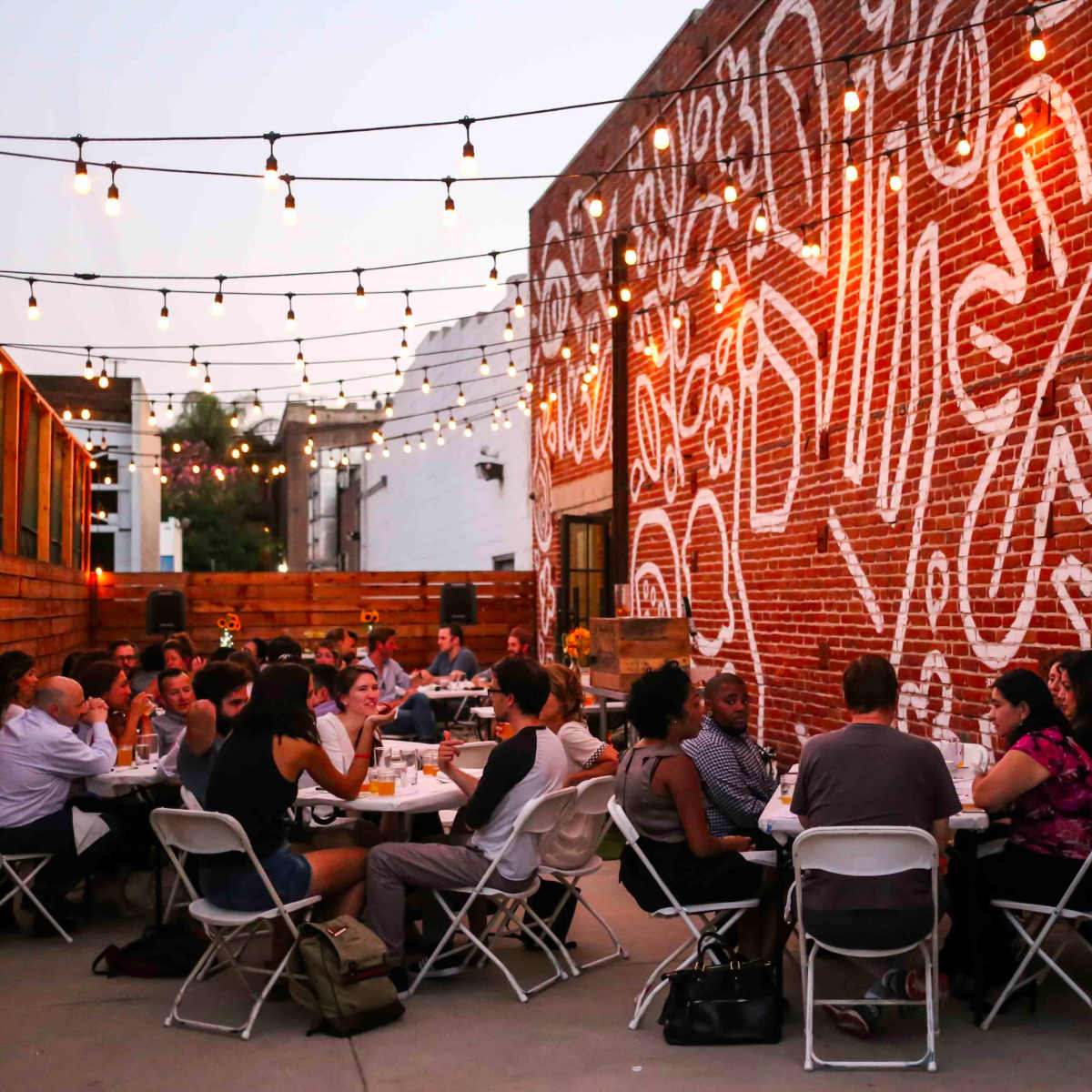 A group of people sits at tables in an outdoor dining area, surrounded by string lights and a mural on a brick wall.