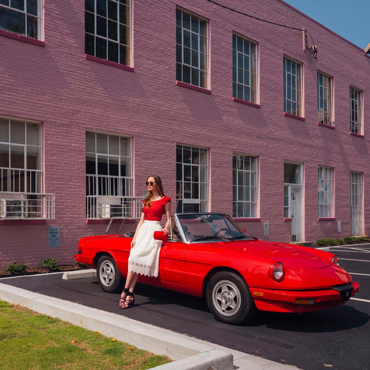 A woman in a red top and white skirt stands next to a red convertible parked in front of a pink building with large windows.