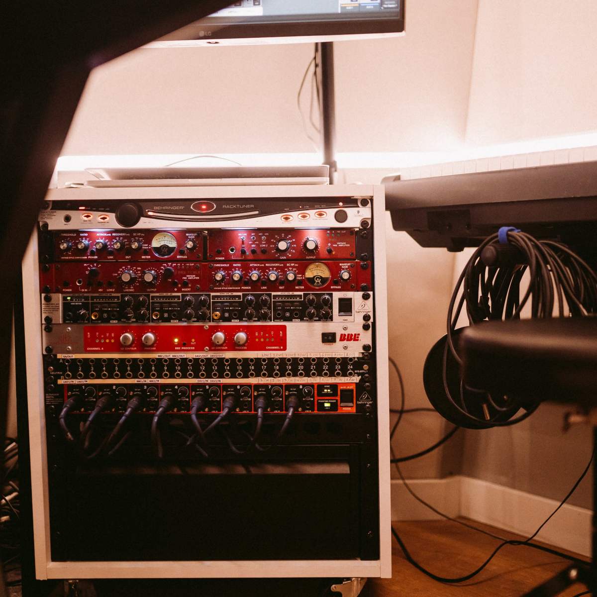 A rack of audio equipment with various control knobs and lights is positioned next to a keyboard and tangled cables in a studio setting.