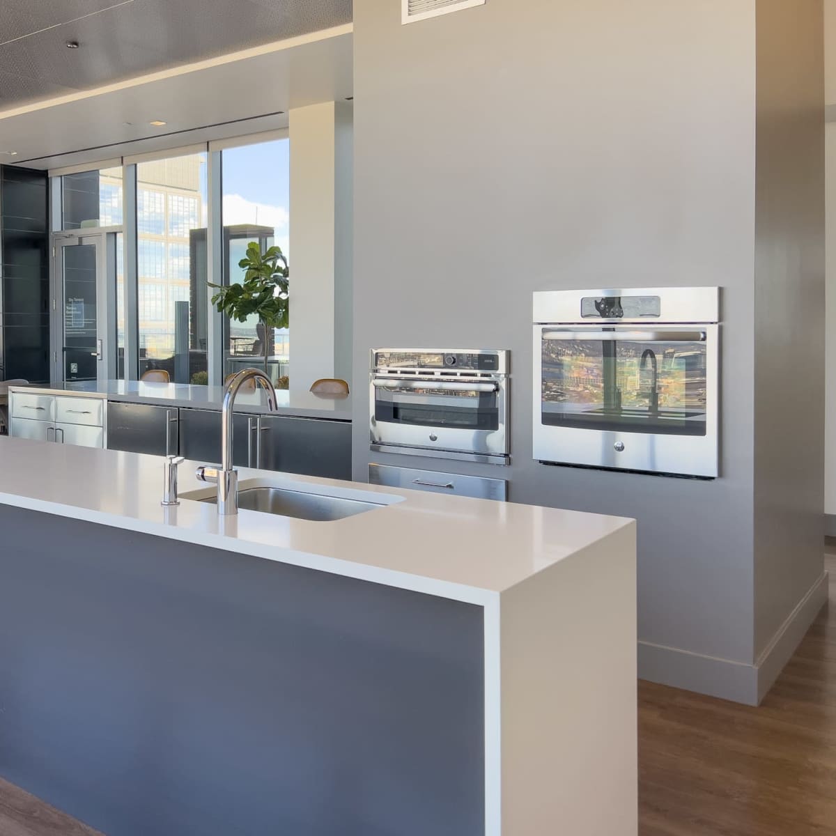 A modern kitchen with stainless steel ovens mounted on a gray wall and a white countertop with a sink.