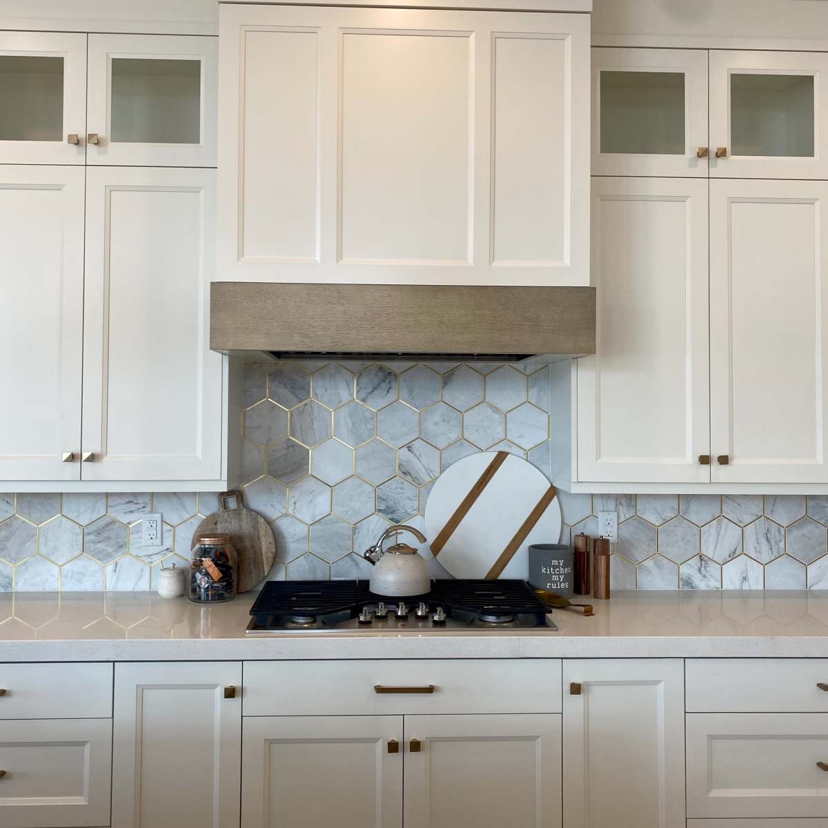 A modern kitchen with white cabinetry, a marble backsplash featuring hexagonal tiles, and a gas cooktop beneath a vent hood.